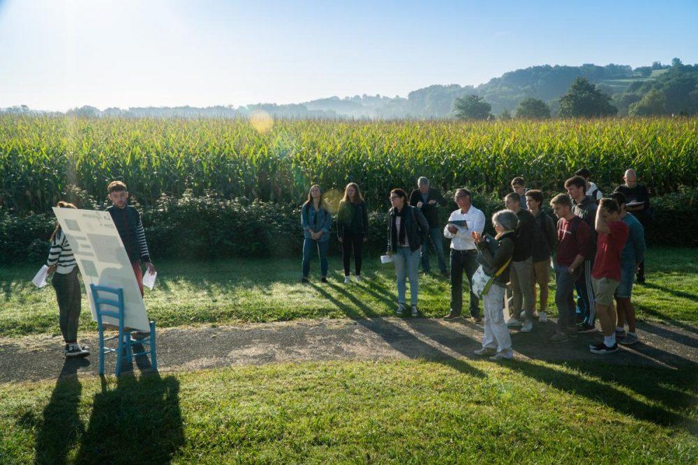 Un étudiant et une étudiante montrent des croquis d'aménagement en bordure du terrain du Foyer, devant les champs de maïs voisins et les collines environnantes.